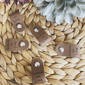 May include: A collection of brown leather tags with silver rivets, each inscribed with "Handmade with Love" in black script. The tags are arranged on a woven, light brown surface, with decorative floral elements in the background.