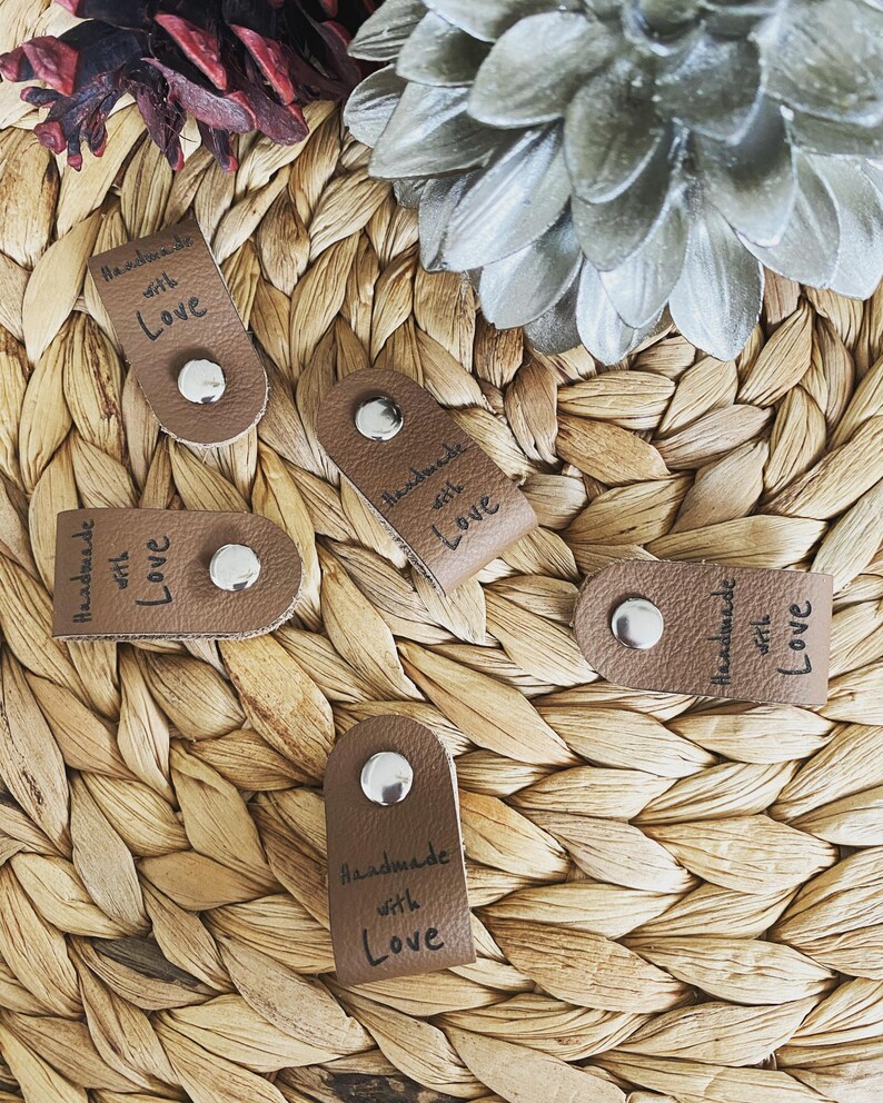 May include: A collection of brown leather tags with silver rivets, each inscribed with "Handmade with Love" in black script. The tags are arranged on a woven, light brown surface, with decorative floral elements in the background.
