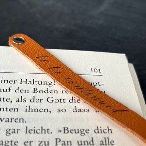 Peut inclure: Un marque-page en cuir beige &agrave; la surface textur&eacute;e, dot&eacute; d'un rivet m&eacute;tallique argent&eacute; sur le dessus. Le marque-page porte l'inscription cursive "le le continue" en encre noire et est pos&eacute; en diagonale sur une page de livre imprim&eacute;e.