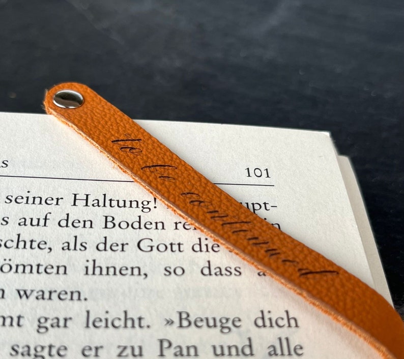 Peut inclure: Un marque-page en cuir beige &agrave; la surface textur&eacute;e, dot&eacute; d'un rivet m&eacute;tallique argent&eacute; sur le dessus. Le marque-page porte l'inscription cursive "le le continue" en encre noire et est pos&eacute; en diagonale sur une page de livre imprim&eacute;e.