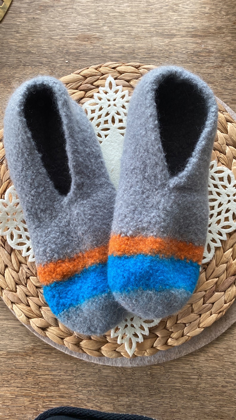 May include: A pair of gray wool slippers with orange and blue stripes. The slippers are on a woven mat.