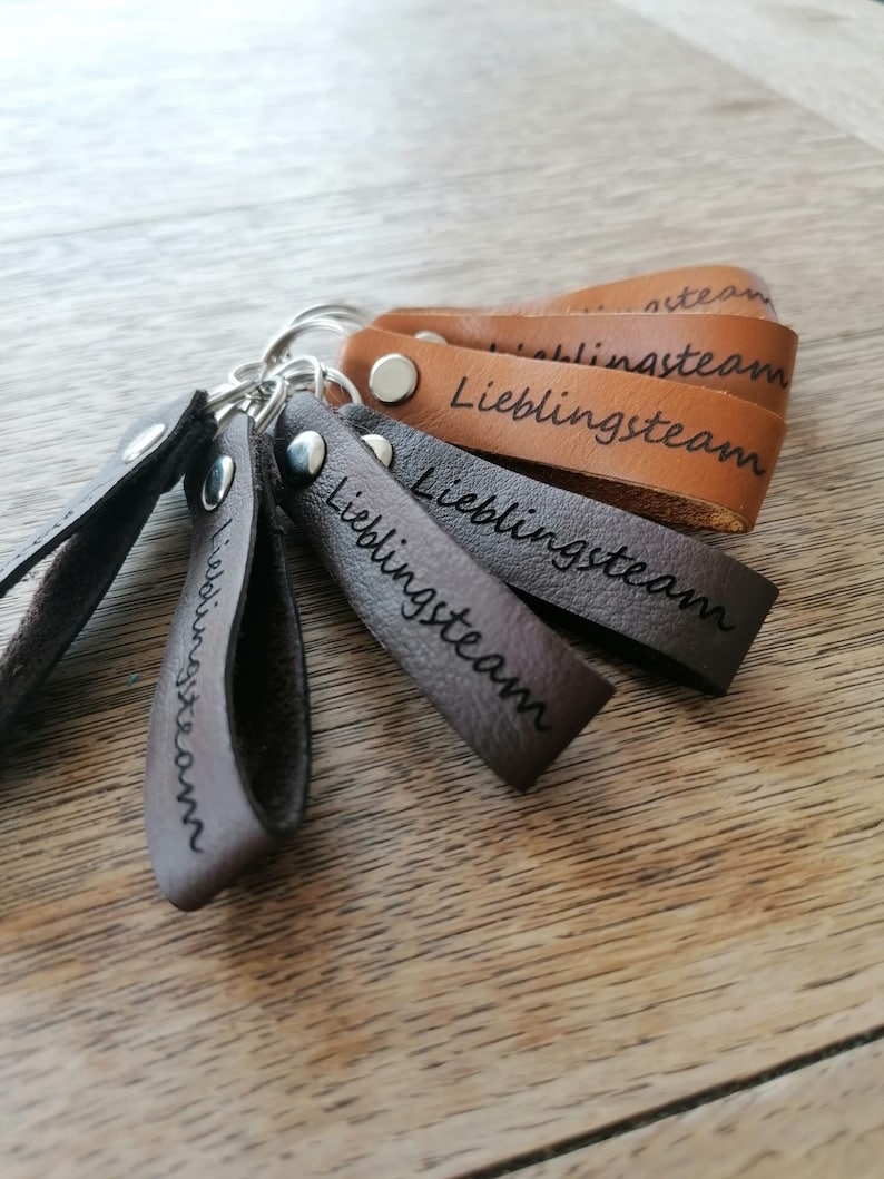 May include: Five brown leather keychains with silver key rings. Each keychain has the word "Lieblingssteam" engraved on it.