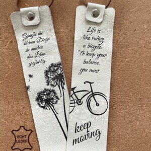 May include: Two white leather bookmarks with black ink. One bookmark features a dandelion design and the text "Genie&szlig;e die kleinen Dinge. Sie machen das Leben gro&szlig;artig." The other bookmark features a bicycle design and the text "Life is like riding a bicycle. To keep your balance, you must keep moving."