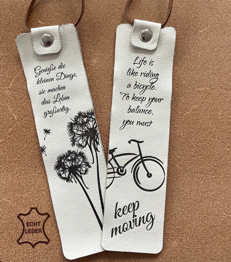 May include: Two white leather bookmarks with black ink. One bookmark features a dandelion design and the text "Genie&szlig;e die kleinen Dinge. Sie machen das Leben gro&szlig;artig." The other bookmark features a bicycle design and the text "Life is like riding a bicycle. To keep your balance, you must keep moving."