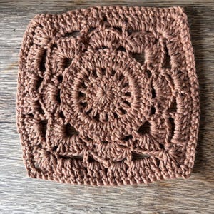 May include: A square crochet motif in a solid dusty rose color. The design features a circular center with radiating stitches, transitioning into a square shape with openwork patterns and shell-like clusters along the edges and corners.