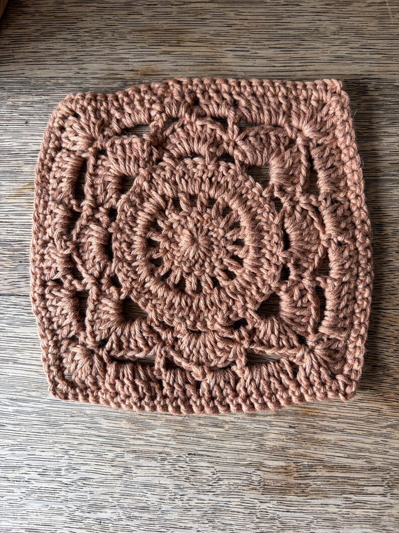 May include: A square crochet motif in a solid dusty rose color. The design features a circular center with radiating stitches, transitioning into a square shape with openwork patterns and shell-like clusters along the edges and corners.