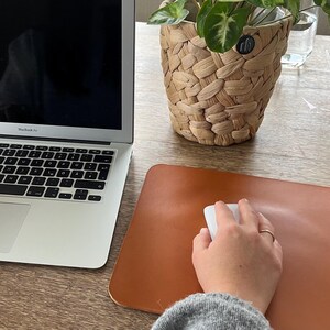 May include: A person is working at a desk with a laptop computer and a brown leather mouse pad. A white wireless mouse is on the pad. The person's hand is on the mouse.