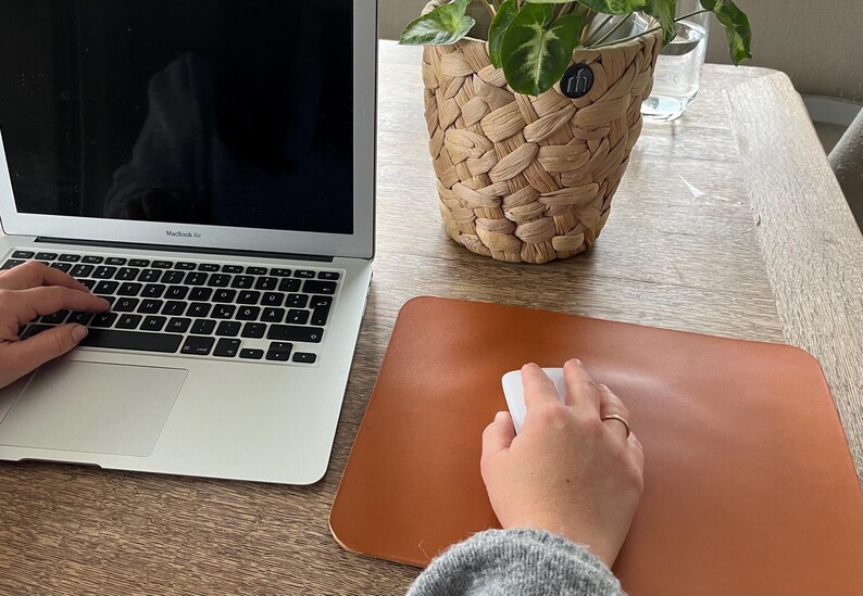 May include: A person is working at a desk with a laptop computer and a brown leather mouse pad. A white wireless mouse is on the pad. The person's hand is on the mouse.