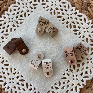 May include: A collection of small, leather tags in various colors, each with unique designs and text. Some tags feature illustrations of a lamb, a garlic clove, and the phrase "C'est la VIE!". The tags are fastened with metal snaps.