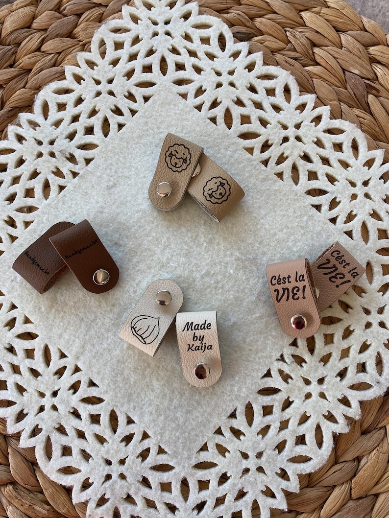 May include: A collection of small, leather tags in various colors, each with unique designs and text. Some tags feature illustrations of a lamb, a garlic clove, and the phrase "C'est la VIE!". The tags are fastened with metal snaps.
