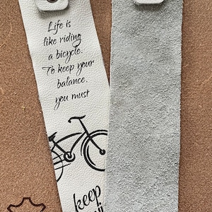 May include: Two white leather bookmarks with a black bicycle illustration and text that reads "Life is like riding a bicycle. To keep your balance, you must keep moving." One bookmark has a brown leather patch with the text "ECHT LEDER".