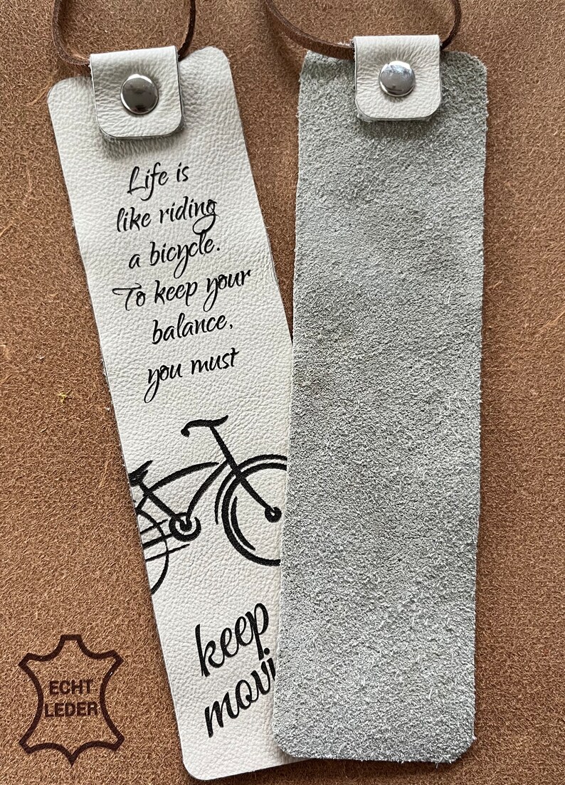 May include: Two white leather bookmarks with a black bicycle illustration and text that reads "Life is like riding a bicycle. To keep your balance, you must keep moving." One bookmark has a brown leather patch with the text "ECHT LEDER".