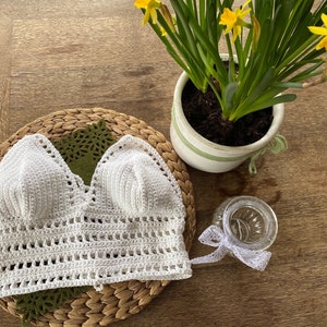 May include: White crochet top with a v-neckline and a delicate crocheted pattern. The top is laid flat on a woven surface.