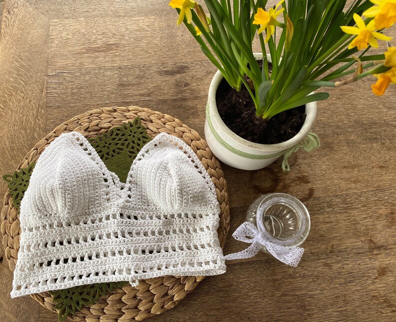 May include: White crochet top with a v-neckline and a delicate crocheted pattern. The top is laid flat on a woven surface.