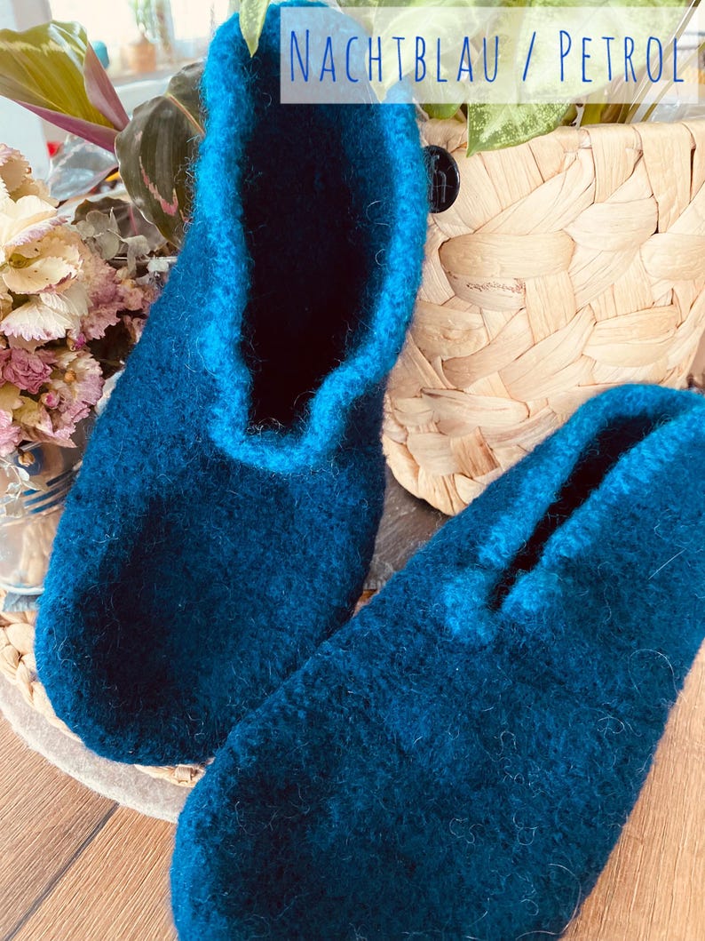 May include: A pair of dark blue wool slippers with a contrasting blue trim. The slippers are on a wooden surface and the text "NACHTBLAU / PETROL" is visible in the background.