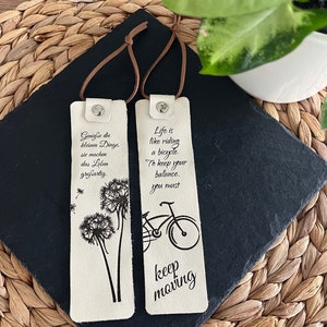 May include: Two white leather bookmarks with black printed text and images. The left bookmark features a dandelion and the text "Geniesse die kleinen Dinge, sie machen das Leben gro&szlig;artig." The right bookmark features a bicycle and the text "Life is like riding a bicycle. To keep your balance, you must keep moving."