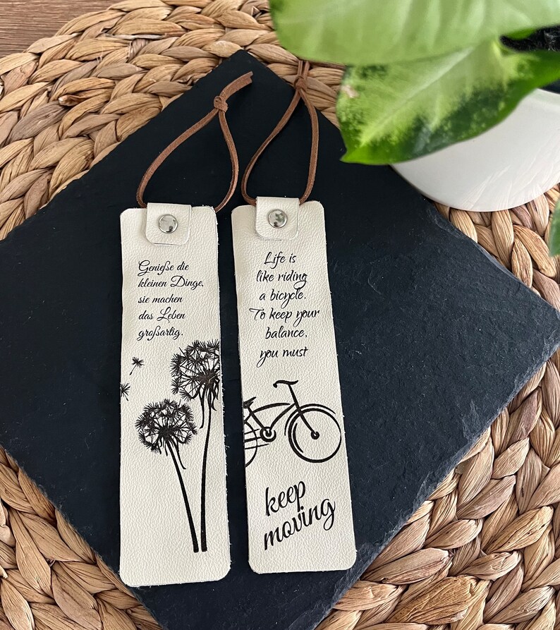 May include: Two white leather bookmarks with black printed text and images. The left bookmark features a dandelion and the text "Geniesse die kleinen Dinge, sie machen das Leben gro&szlig;artig." The right bookmark features a bicycle and the text "Life is like riding a bicycle. To keep your balance, you must keep moving."