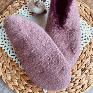 May include: A pair of soft, fuzzy slippers in a light purple color with a dark purple trim around the opening. The slippers are sitting on a woven wicker mat.