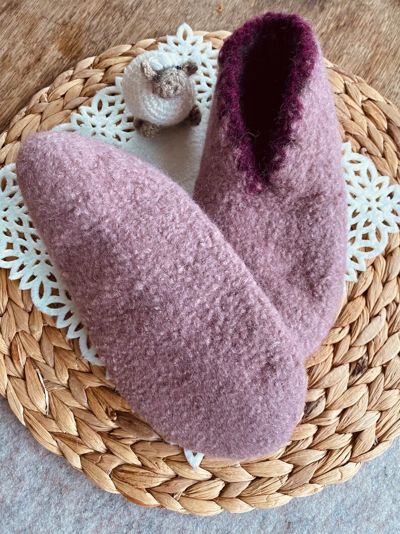 May include: A pair of soft, fuzzy slippers in a light purple color with a dark purple trim around the opening. The slippers are sitting on a woven wicker mat.