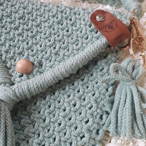 May include: Close-up of a light green macrame bag with a woven pattern. The bag features a wooden bead, a leather tag with a design, and a tassel. The bag has a braided strap and a gold-colored clasp.
