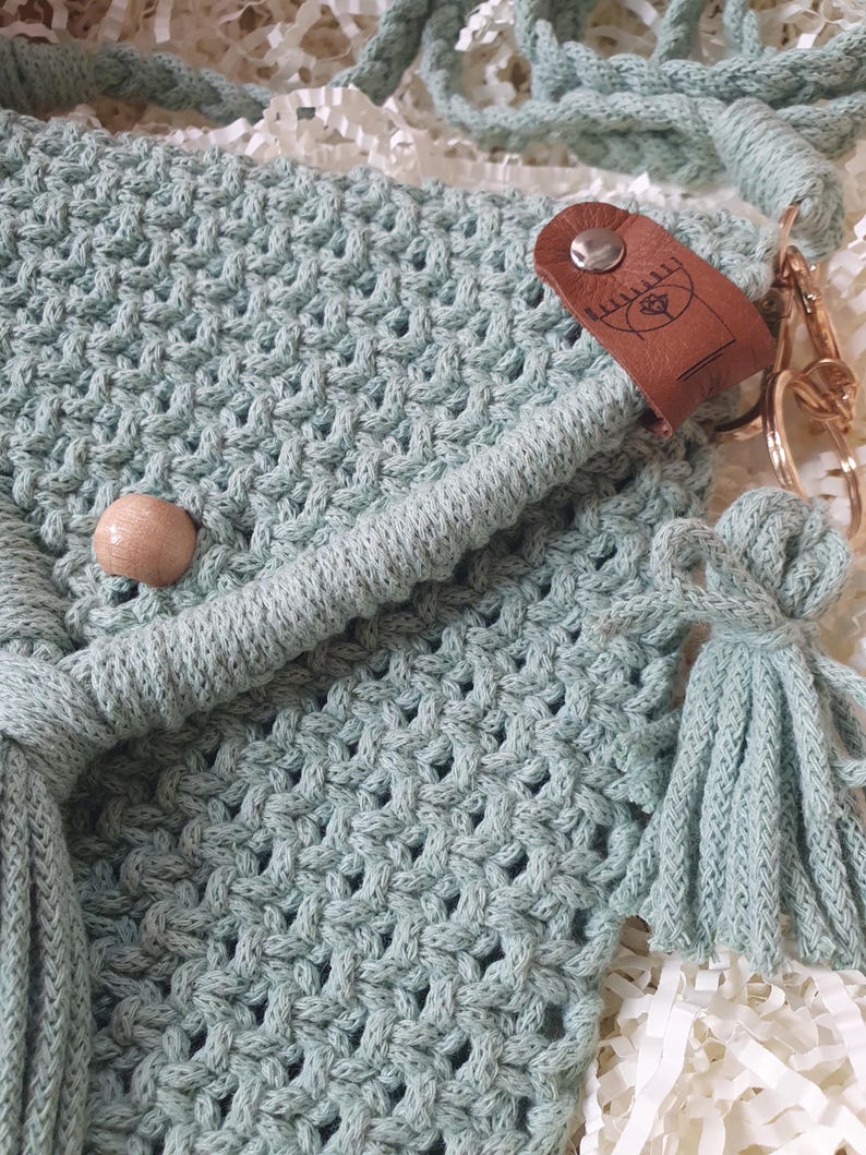 May include: Close-up of a light green macrame bag with a woven pattern. The bag features a wooden bead, a leather tag with a design, and a tassel. The bag has a braided strap and a gold-colored clasp.