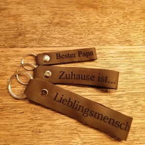 May include: Three brown leather keychains with silver key rings. The keychains have the following text engraved on them: "Bester Papa", "Zuhause ist...", and "Lieblingsmensch".