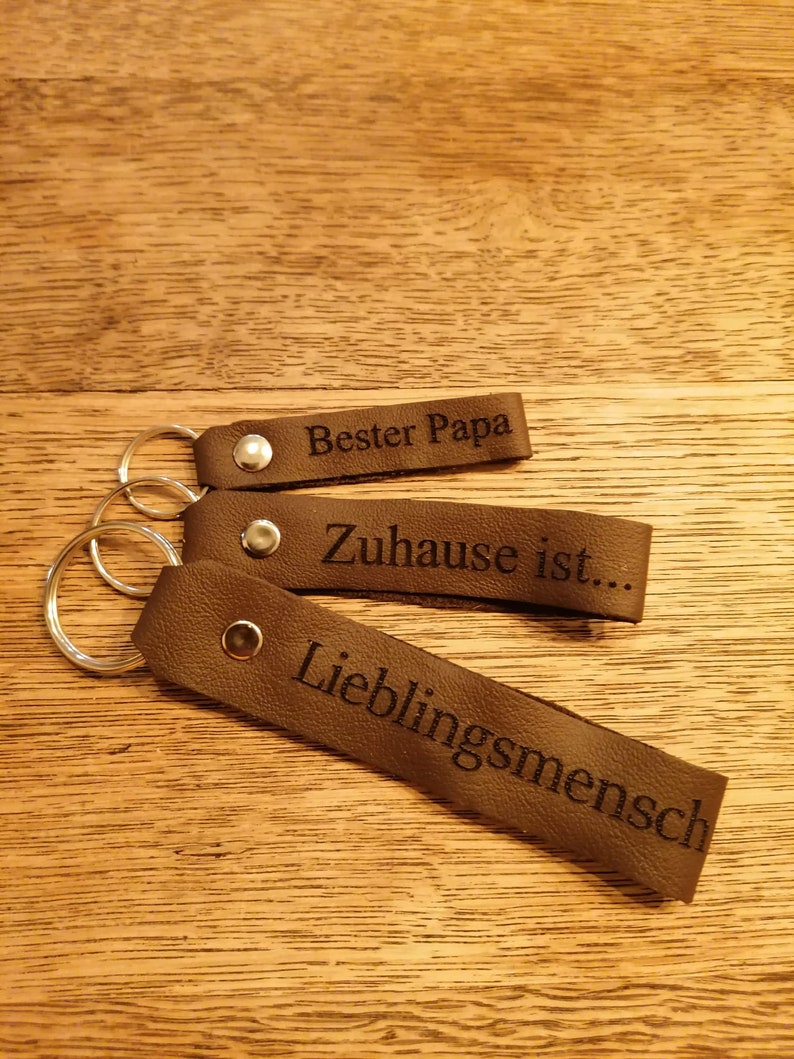 May include: Three brown leather keychains with silver key rings. The keychains have the following text engraved on them: "Bester Papa", "Zuhause ist...", and "Lieblingsmensch".