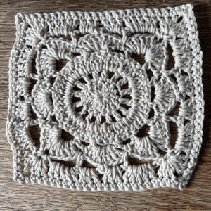 May include: A square crochet motif in a light beige color featuring a circular center with radiating stitches, surrounded by a series of shell-like patterns that transition the circular design into a square shape with openwork spaces throughout.