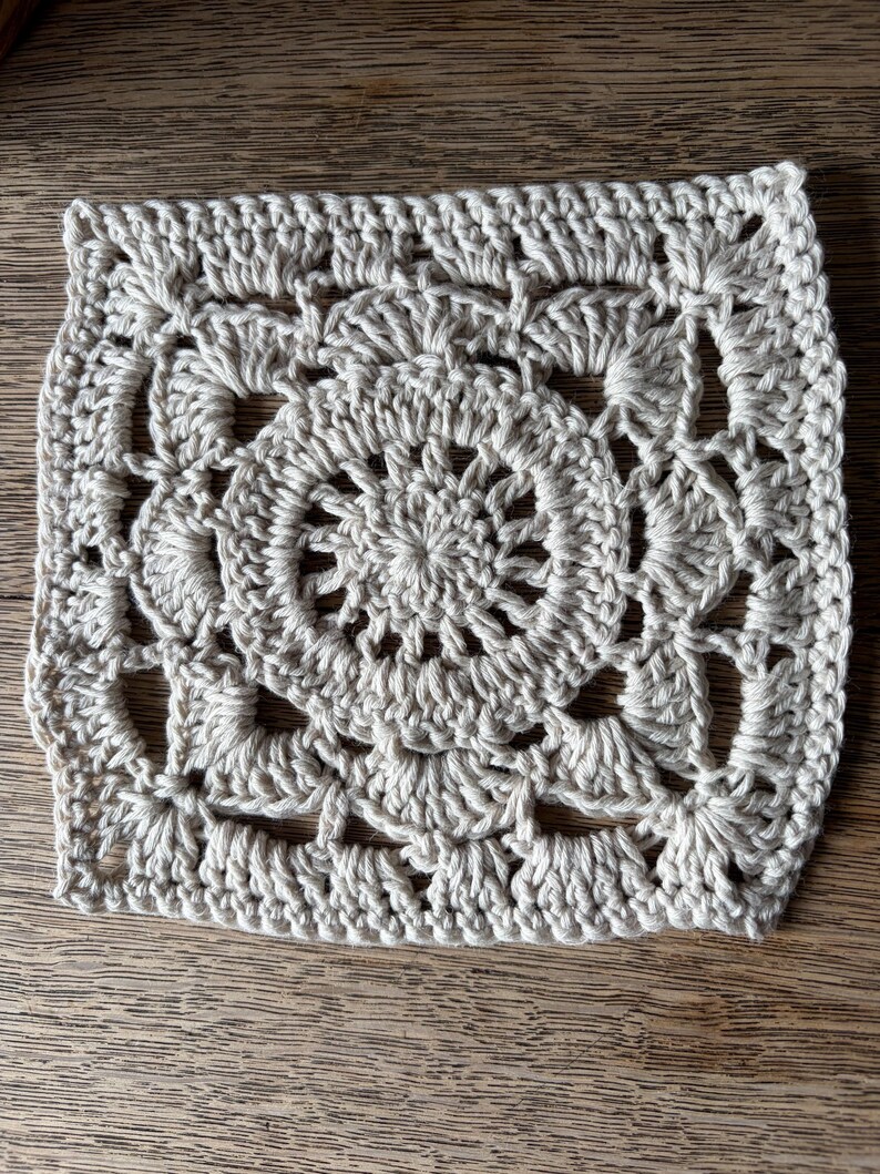 May include: A square crochet motif in a light beige color featuring a circular center with radiating stitches, surrounded by a series of shell-like patterns that transition the circular design into a square shape with openwork spaces throughout.