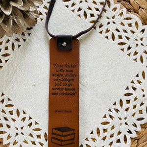 May include: Brown leather bookmark with a quote by Francis Bacon: "Einge B&uuml;cher sollte man kosten, andere verschlingen und einige wenige kauen und verdauen". The bookmark features a stack of books in the bottom corner.