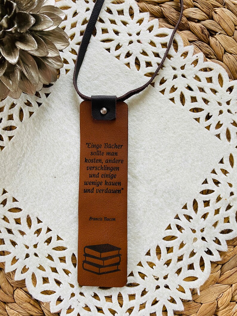 May include: Brown leather bookmark with a quote by Francis Bacon: "Einge B&uuml;cher sollte man kosten, andere verschlingen und einige wenige kauen und verdauen". The bookmark features a stack of books in the bottom corner.
