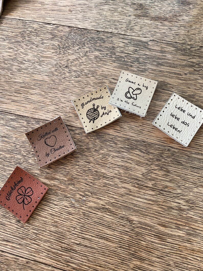 May include: Five different leather labels with various designs and text. The labels are beige and brown. The text on the labels includes "Knitted with love by Christine", "Handmade by Anja", "Gimme a hug I'm the future", "Gl&uuml;ckskind" and "Lebe und liebe das Leben!"