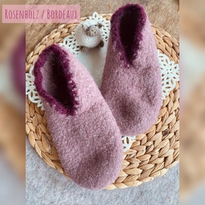 May include: Two pairs of soft, pink knitted slippers with a burgundy trim. The slippers are sitting on a woven wicker basket.