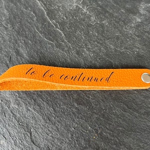 Peut inclure: Un marque-page en cuir orange avec un rivet en m&eacute;tal argent&eacute; &agrave; une extr&eacute;mit&eacute; et le texte cursif noir "to be continued..." imprim&eacute; au centre de la lani&egrave;re. Le marque-page pr&eacute;sente une texture lisse et un bord pli&eacute; l&eacute;g&egrave;rement incurv&eacute;.