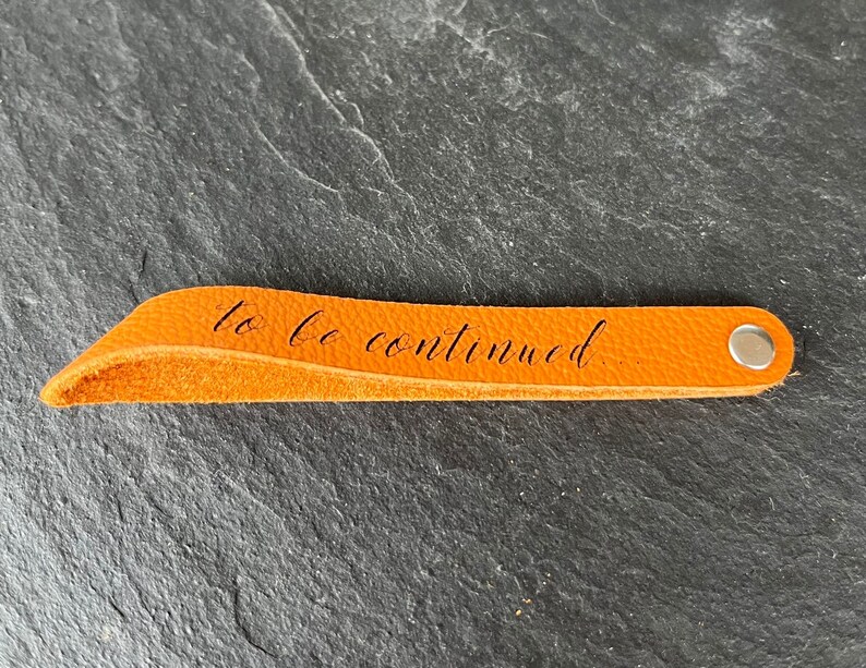 Peut inclure: Un marque-page en cuir orange avec un rivet en m&eacute;tal argent&eacute; &agrave; une extr&eacute;mit&eacute; et le texte cursif noir "to be continued..." imprim&eacute; au centre de la lani&egrave;re. Le marque-page pr&eacute;sente une texture lisse et un bord pli&eacute; l&eacute;g&egrave;rement incurv&eacute;.
