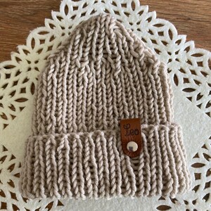 May include: A beige knit beanie with a folded brim. The hat features a leather tag with the name "Leo" and a button. The beanie is made of thick yarn and has a ribbed texture.