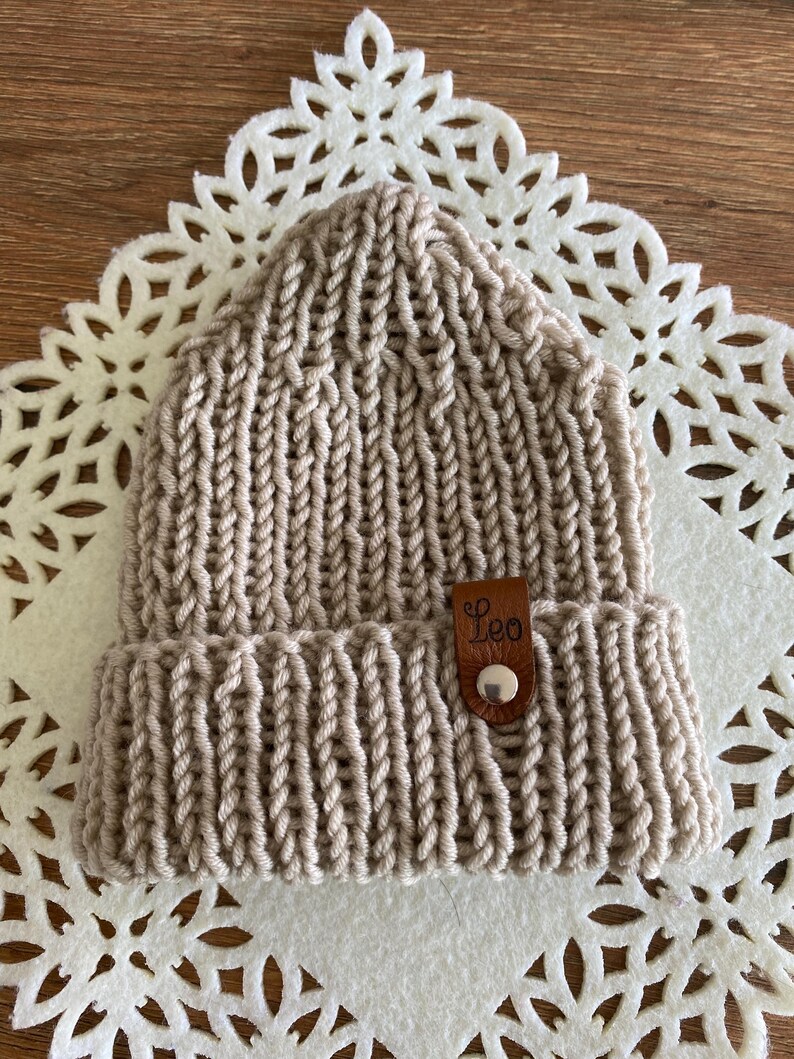 May include: A beige knit beanie with a folded brim. The hat features a leather tag with the name "Leo" and a button. The beanie is made of thick yarn and has a ribbed texture.