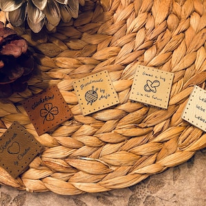 May include: Five small, square, brown leather tags with black text and illustrations. The tags are on a woven brown background. The text on the tags reads: "Knitted with love by Christine", "Gl&uuml;ckskind", "Handmade by Anja", "Gimme a bag I'm the future", and "Lebe und liebe das Leben!"