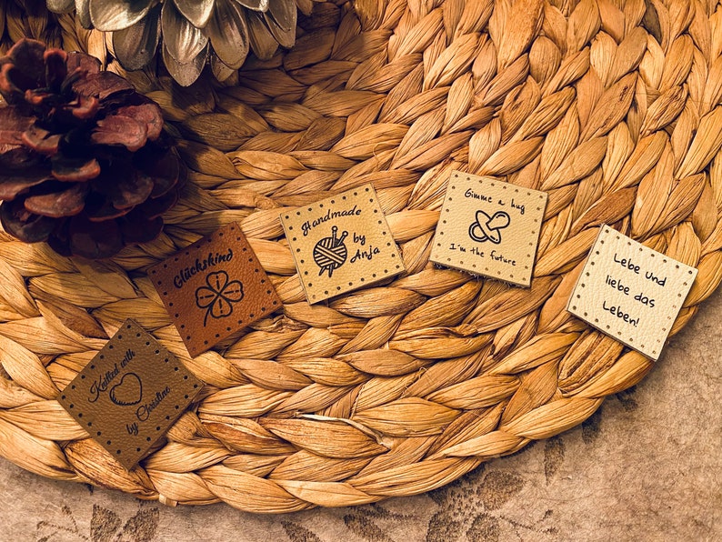 May include: Five small, square, brown leather tags with black text and illustrations. The tags are on a woven brown background. The text on the tags reads: "Knitted with love by Christine", "Gl&uuml;ckskind", "Handmade by Anja", "Gimme a bag I'm the future", and "Lebe und liebe das Leben!"