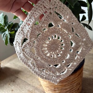 May include: A square-shaped off-white crochet piece featuring a circular mandala-style pattern in the center that transitions into a square border with intricate stitch work and open spaces throughout the design, crafted from light-colored yarn.