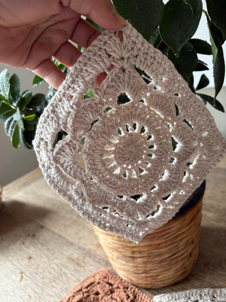 May include: A square-shaped off-white crochet piece featuring a circular mandala-style pattern in the center that transitions into a square border with intricate stitch work and open spaces throughout the design, crafted from light-colored yarn.