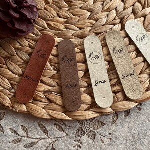 May include: Five leather tags in shades of brown, beige, and cream, each with a circular logo and text. The tags are arranged on a woven surface. The text on the tags includes "Marone", "Nuss", "Grau", "Sand", and "Creme".