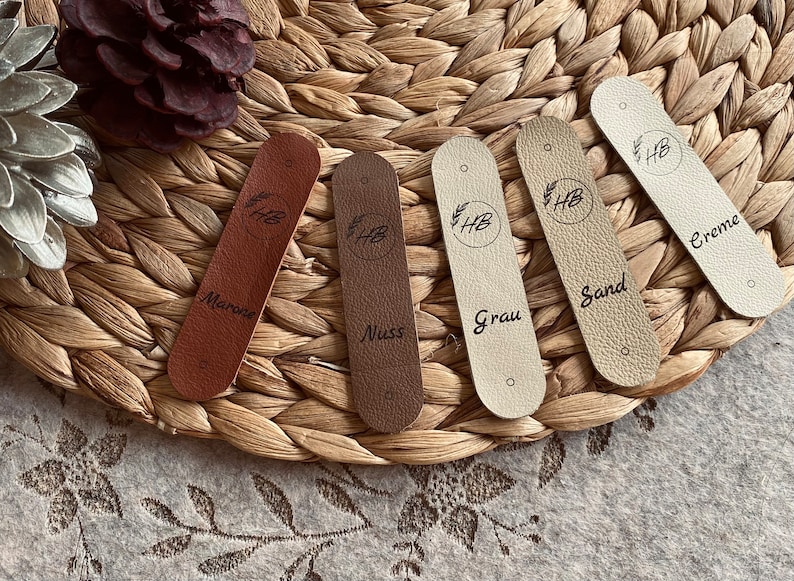 May include: Five leather tags in shades of brown, beige, and cream, each with a circular logo and text. The tags are arranged on a woven surface. The text on the tags includes "Marone", "Nuss", "Grau", "Sand", and "Creme".