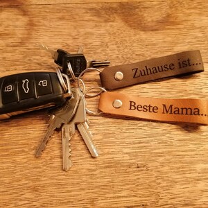 May include: A set of two leather keychains with engraved text. The first keychain is brown and reads "Zuhause ist..." The second keychain is tan and reads "Beste Mama..."