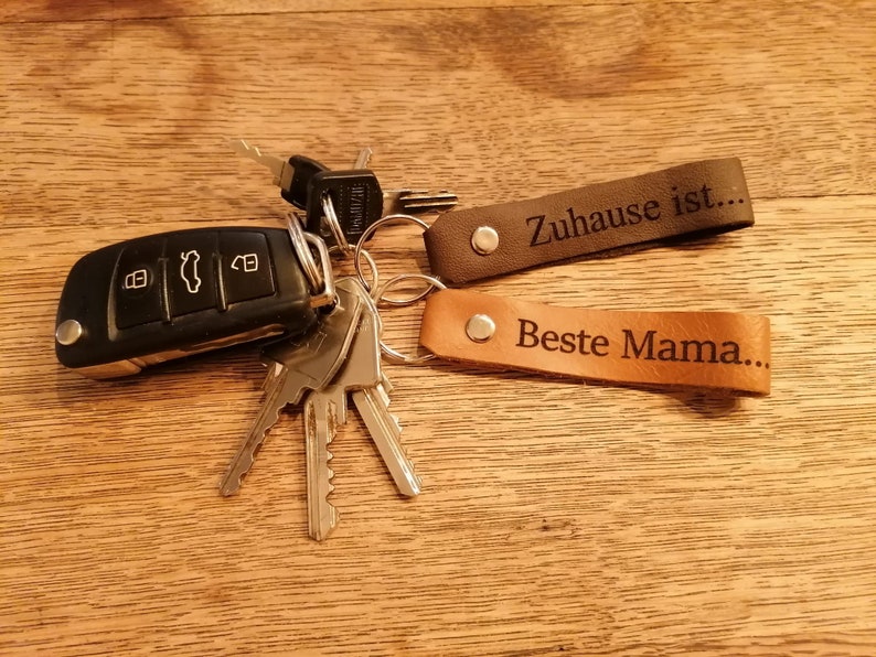 May include: A set of two leather keychains with engraved text. The first keychain is brown and reads "Zuhause ist..." The second keychain is tan and reads "Beste Mama..."