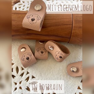 May include: Tan leather dog-themed accessories with a metal snap closure. The accessories feature a dog face design and the text "MIT EIGENEM LOGO" and "IN ROTBRAUN".