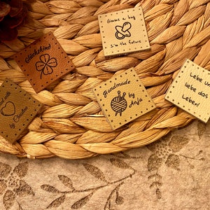 May include: Five brown leather labels with different text and images. The labels include "Knitted with love by Christine", "Gl&uuml;ckskind", "Gimme a hug I'm the future", "Handmade by Anja" with a yarn ball and knitting needles, and "Lebe und liebe das Leben!"