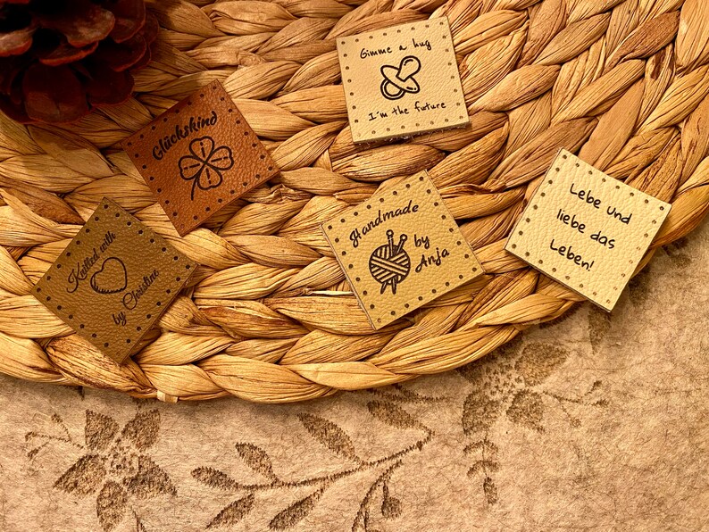 May include: Five brown leather labels with different text and images. The labels include "Knitted with love by Christine", "Gl&uuml;ckskind", "Gimme a hug I'm the future", "Handmade by Anja" with a yarn ball and knitting needles, and "Lebe und liebe das Leben!"