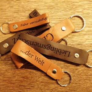 May include: Five leather keychains with metal rings. The keychains are brown and black and have different engraved messages, including "Fahr vorsichtig!" and "...der Welt".