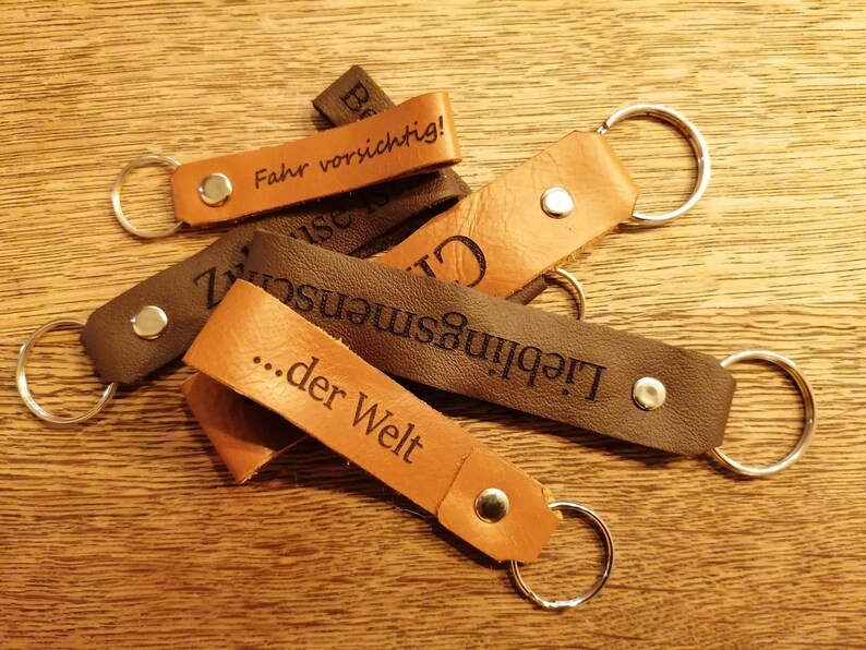 May include: Five leather keychains with metal rings. The keychains are brown and black and have different engraved messages, including "Fahr vorsichtig!" and "...der Welt".